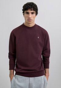 Bordeaux sweatshirt med ribbet tekstur, rund halsudskæring og en lille hvid logoaccent på brystet, båret med grå bukser med elastisk talje.