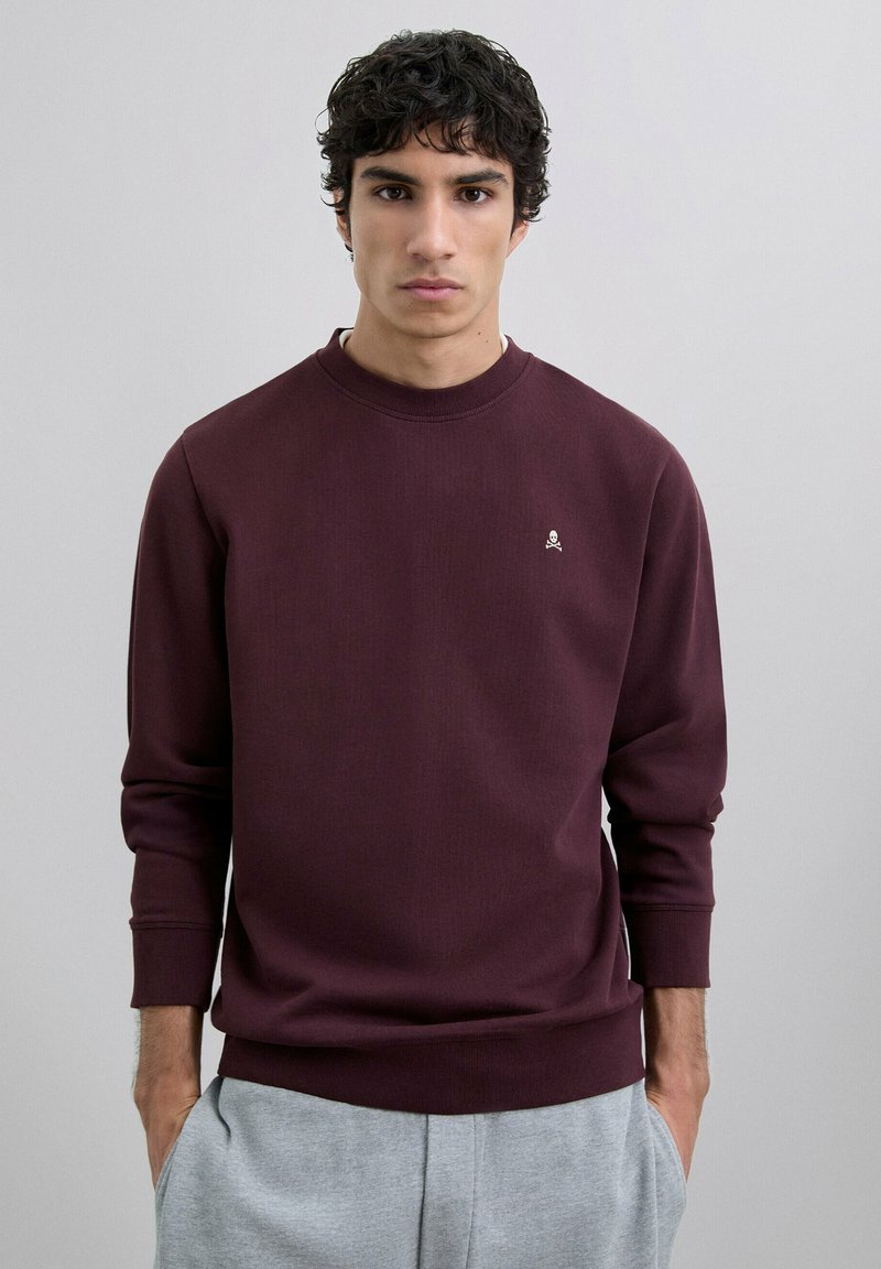 Bordeaux sweatshirt med ribbet tekstur, rund halsudskæring og en lille hvid logoaccent på brystet, båret med grå bukser med elastisk talje.