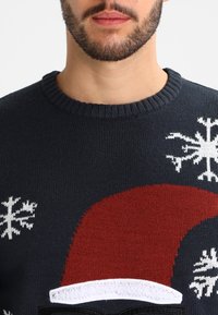 Homme avec barbe portant un pull en tricot foncé décoré d'un bonnet de Père Noël rouge et de flocons de neige blancs.