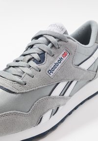 Gros plan d'une basket Reebok Classic grise et blanche avec des accents en daim, des lacets et un petit patch drapeau Union Jack sur le côté.
