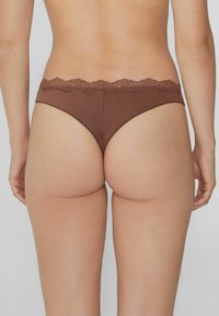 Rückansicht einer Person, die braune, mit Spitze verzierten Bikini-Unterwäsche trägt, vor einem einfarbigen Hintergrund.