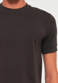 Nærbillede af en person iført en ensfarvet sort T-shirt med korte ærmer, ribbet rund halsudskæring og ribbede ærmekanter.