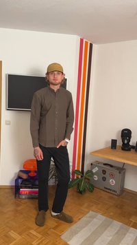 Tmavohnědá košile na knoflíky, černé kalhoty a hnědé nazouvací boty. Krátké světlé vlasy pod béžovou čepicí. Stojí v moderním interiéru.