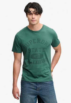 Ung mand med mørkt hår iført en grøn Superdry State New York t-shirt og blå jeans, stående mod en ensartet lys baggrund.