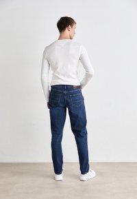 Garās piedurknes balta krekla saderība ar tumši ziliem džinsiem. Džinsiem ir divas aizmugures kabatas un brūna birka pie jostasvietas. Baltas sporta apavi.