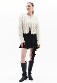 Giacca bianca con frange dal design corto, abbinata a una mini gonna nera a righe con orlo arricciato e stivali neri alti fino al ginocchio.