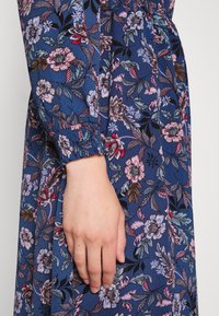 Main reposant contre une robe bleue avec un motif floral rose, violet et marron et un poignet froncé sur une manche longue.