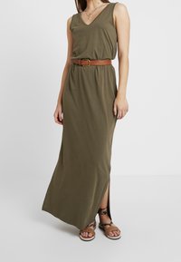 Oliwkowa, bezramienna sukienka maxi z dekoltem w kształcie V, z paskiem w talii i bocznymi rozporkami. W zestawie z płaskimi sandałami na kostkę.