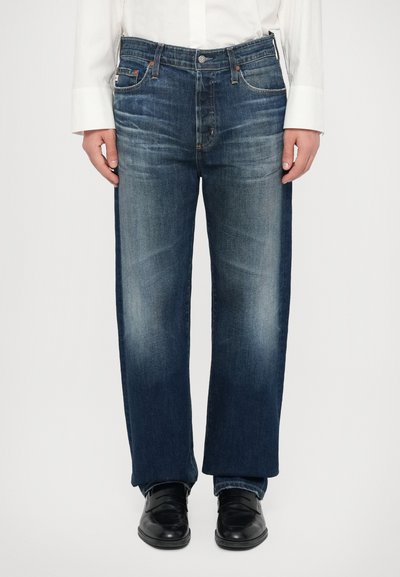 Jean en denim bleu avec une coupe décontractée, présentant un délavage près des genoux et des accents de couture orange. Portés avec des chaussures noires.