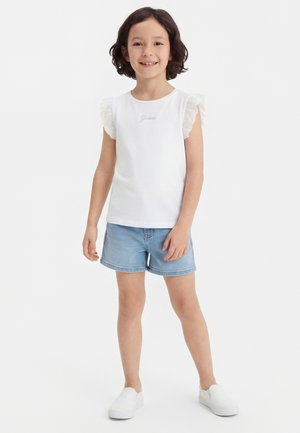 Fată cu păr scurt și creț zâmbind, purtând un top alb fără mâneci cu volane, pantaloni scurți din denim albastru deschis și pantofi albi slip-on.