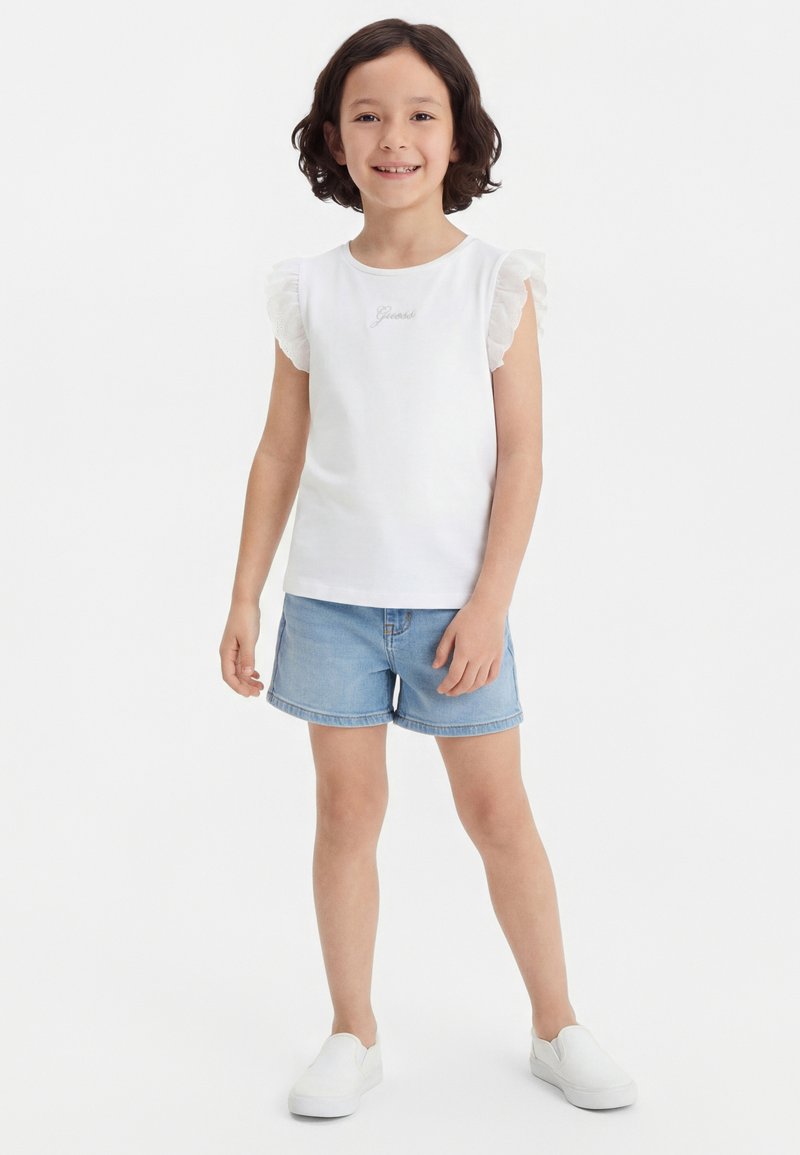 Κορίτσι με κοντά σγουρά μαλλιά που χαμογελά, φοράει λευκή αμάνικη μπλούζα με βολάν, ανοιχτό μπλε τζιν σορτς και λευκά παπούτσια χωρίς κορδόνια.