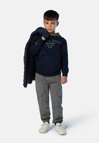 Sudadera azul marino con bolsillo frontal y texto, pantalones cargo grises con bolsillos laterales, sosteniendo una chaqueta acolchada oscura, zapatillas blancas.