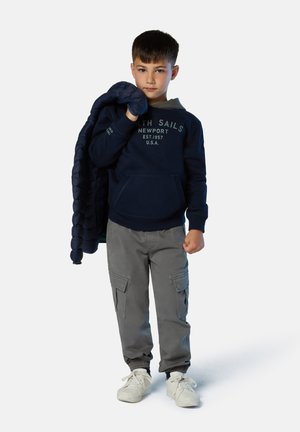 Sudadera azul marino con bolsillo frontal y texto, pantalones cargo grises con bolsillos laterales, sosteniendo una chaqueta acolchada oscura, zapatillas blancas.