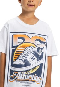 Bílý bavlněný tričko s grafickým potiskem tenisky a textem "DC Athletics" v modré a oranžové barvě, s kulatým pozadím paprsků slunce.
