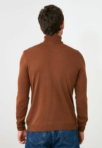Maglione a collo alto marrone realizzato in materiale a coste, design aderente, maniche lunghe e colletto avvolgente. Indossato con jeans in denim blu.
