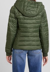 Olivgrüne Steppjacke mit Kapuze, mit gestepptem Design, gerippten Bündchen und seitlichen Nähten für eine bessere Passform. Getragen mit ausgebleichten blauen Jeans.