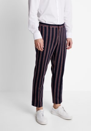 Mann trägt marineblaue Hose mit roten und weißen vertikalen Streifen, weißes Hemd und weiße Turnschuhe, steht vor einfarbigem Hintergrund.
