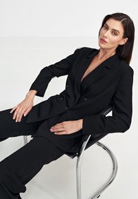 Czarna, dopasowana marynarka z głębokim dekoltem w kształcie litery V i podwójnym zapięciem, zestawiona z dopasowanymi spodniami, na minimalistycznym tle.