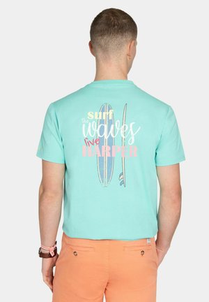 Hombre vistiendo una camiseta turquesa con tablas de surf y el texto "Surf the waves live Harper", combinado con pantalones cortos naranjas, visto desde atrás.