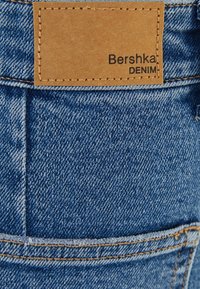 Modrá džínová tkanina obsahuje zadní kapsu s koženou značkou v barvě tan. Na značce je černým písmem napsáno "Bershka DENIM". Viditelné jsou švy.