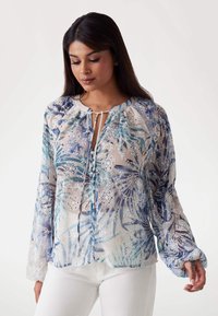 Bluză din material transparent cu imprimeu floral albastru, având un decolteu tip cheie, un sistem de strângere în față și mâneci lungi cu manșete gathered.