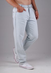 Mann in hellblauen Jeans und weißen Sneakers, steht mit einer Hand in der Tasche vor einem schlichten grauen Hintergrund.