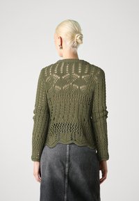 Pull tricoté vert olive avec un motif ajouré, manches longues et ourlet festonné. Porté avec une jupe gris foncé.