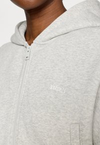 Ανοιχτό γκρι φούτερ με φερμουάρ από απαλό, υφασμάτινο υλικό με υφή. Διαθέτει κουκούλα και τη λέξη «STRONGER» κεντημένη με λευκά γράμματα στο μπροστινό μέρος.