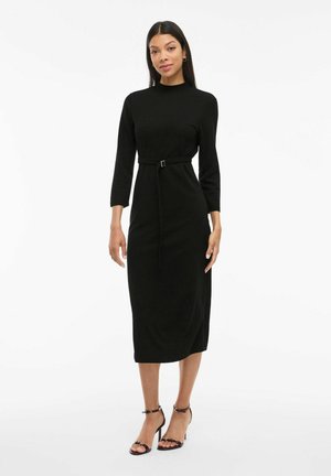 Black, long-sleeve midi dress with a fitted waist and a belt detail. Smooth fabric with a high neckline, complemented by open-toed heels.