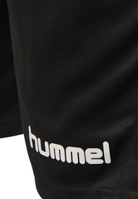 Pantaloni sportivi neri realizzati in tessuto leggero, con il logo bianco "hummel" su un lato e una texture liscia e opaca.