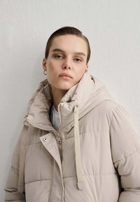 Giacca imbottita beige con collo alto, cappuccio regolabile con cordino, zip sul davanti e trama trapuntata. Tessuto liscio con vestibilità rilassata.