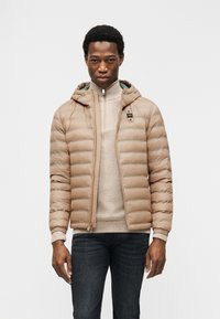 Νεαρός άνδρας φορούντας μπεζ φουσκωτό μπουφάν πάνω από ένα ανοιχτό μπεζ πουλόβερ, συνδυασμένο με σκούρα τζιν, πάνω σε απλό λευκό φόντο.