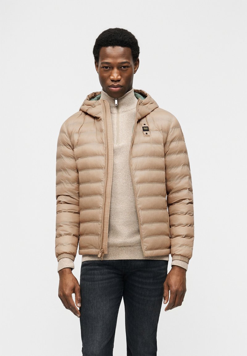 Νεαρός άνδρας φορούντας μπεζ φουσκωτό μπουφάν πάνω από ένα ανοιχτό μπεζ πουλόβερ, συνδυασμένο με σκούρα τζιν, πάνω σε απλό λευκό φόντο.