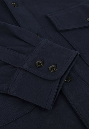 Donkerblauwe langetermijn shirtmanchetten met twee zwarte knopen, gedetailleerde stiksels, en een deel van een borstzak met klep zichtbaar.