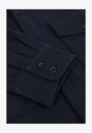 Donkerblauwe langetermijn shirtmanchetten met twee zwarte knopen, gedetailleerde stiksels, en een deel van een borstzak met klep zichtbaar.