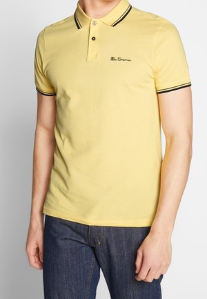 Mann trägt gelbes Ben Sherman-Poloshirt mit schwarzem Kragen- und Ärmelbesatz, kombiniert mit dunkelblauen Jeans, steht vor einfarbigem Hintergrund.
