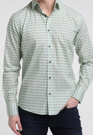 Homme portant une chemise à manches longues boutonnée à motifs verts clairs avec un jean foncé, main dans la poche sur fond uni.