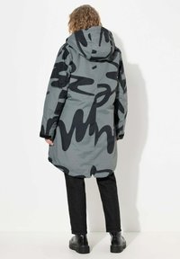 Larga chaqueta gris con patrones abstractos negros, capucha y un corte relajado. Combinada con jeans negros y botas negras gruesas. Textura suave.