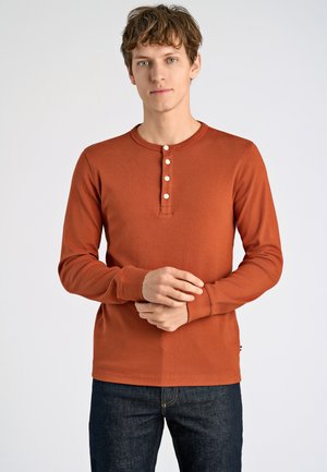 Roestkleurig langetermijn henley overhemd gemaakt van zachte stof, met een ronde halslijn en een vijfknopenbies. Gecombineerd met donkere jeans.