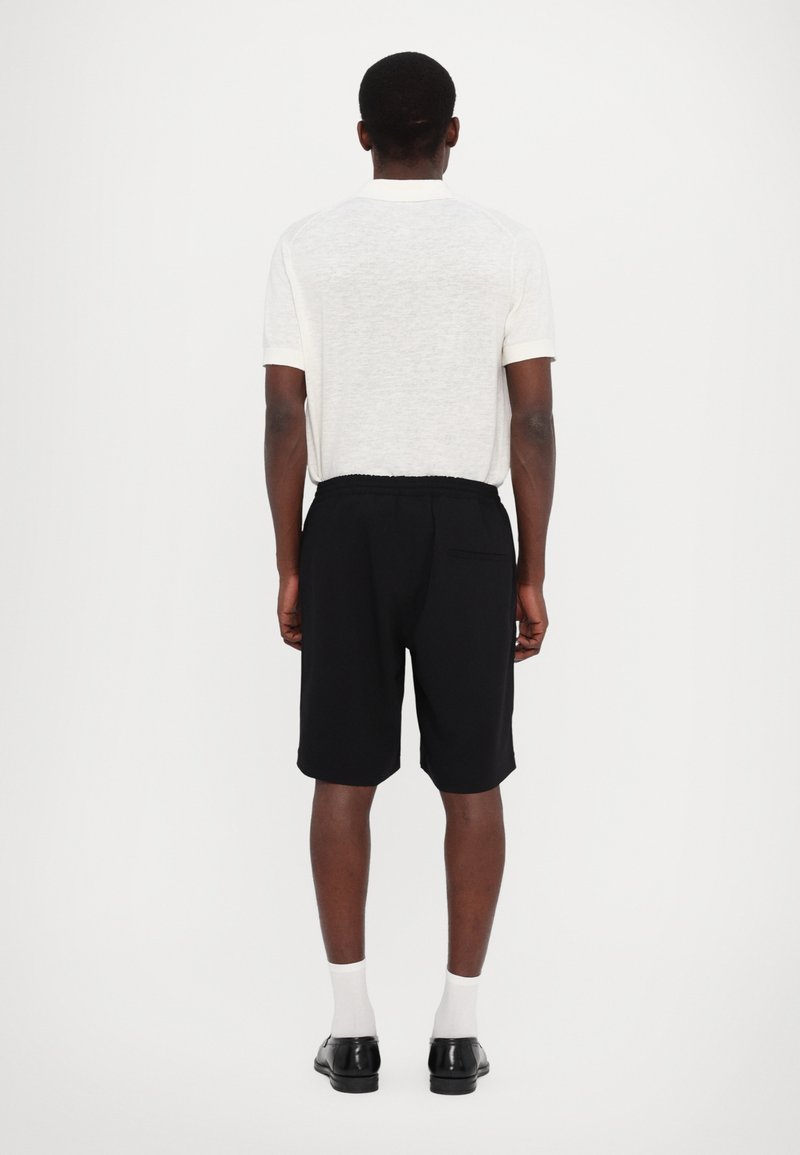 Camicetta bianca in lino a maniche corte con risvolti e pantaloni corti neri con vita elastica. Le scarpe nere e i calzini bianchi completano l'outfit.