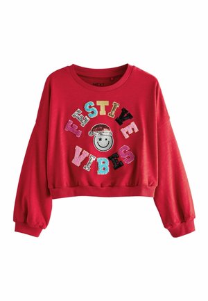 Sweatshirt rouge court à manches longues avec un visage souriant et un texte en sequins épelant "FESTIVE VIBES" dans diverses couleurs et polices.