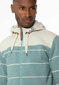 Capuchon sweatshirt in teal en crème, met horizontale witte strepen, een volledige rits, trekkoordaccenten en een merketiket op de kraag.
