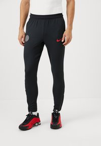 Sorte sportbukser med ribbet tekstur, elastisk talje; har et rødt Nike-logo og en grå cirkulær detalje på venstre lår.
