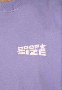 Lys lilla T-skjorte laget av bomull. Har hvit trykt tekst "DROP SIZE" med stjernedetalj. Glatt tekstur, klassisk rund hals.