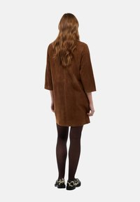 Robe tunique en daim marron avec manches trois-quarts, ourlet arrondi et coupe décontractée. Associée à des collants foncés et des chaussures à motifs.