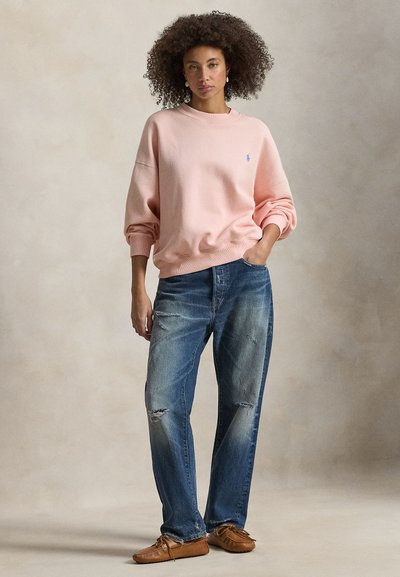 Ανοιχτό ροζ φούτερ με χαλαρή εφαρμογή, ριμπ μανσέτες και μικρό λογότυπο. Συνδυασμένο με σκούρο μπλε τζιν με φθορές και καφέ μοκασίνια.