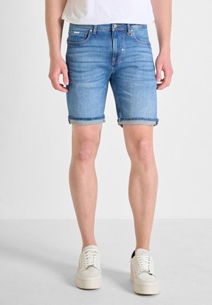 Man draagt blauwe opgerolde denim shorts en witte sneakers, staande op een lichte achtergrond.