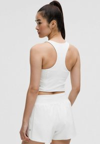 Jeune femme aux cheveux noirs longs en queue de cheval, portant un crop top blanc à dos nageur et un short de sport blanc, tournée vers l'arrière contre un fond uni.