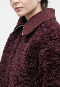 Detailný záber osoby v tmavofialovom kabáte z ovčej vlny s rebrovaným vysokým golierom a viditeľným strieborným zipsom.