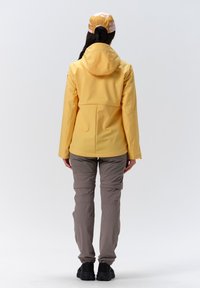 Waterdichte gele jas met capuchon, gecombineerd met lichtgrijze broek en zwarte schoenen. Gladde textuur, praktische uitstraling met elastische manchetten.